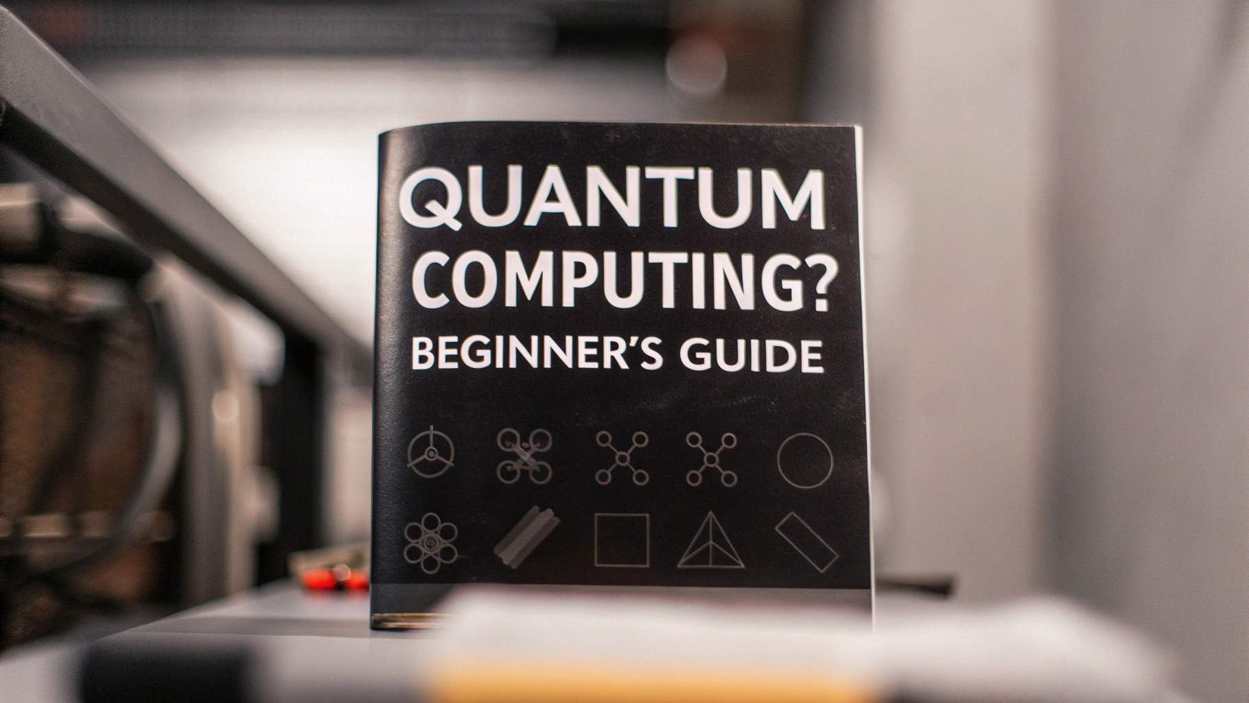 A black booklet titled "Quantum Computing? Beginner's Guide" is standing upright on a metallic surface. The title is printed in bold white capital letters, and the bottom section features various abstract scientific and geometric symbols in grey. The background is softly blurred, highlighting the focus on the booklet.