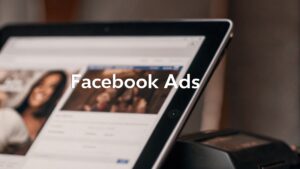 Facebook Ads text overlay on a blurred image of a tablet screen showing Facebook-like profiles and content.